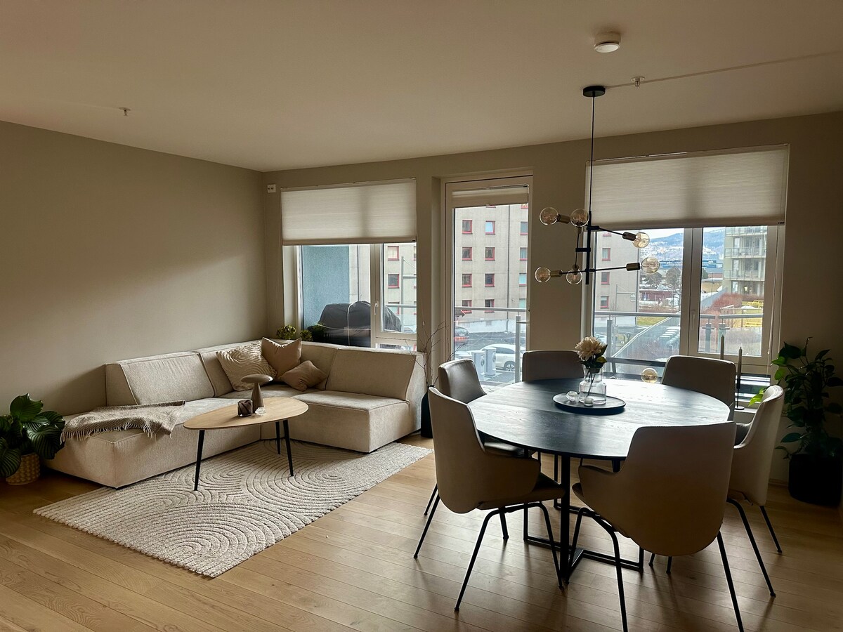 A modern living area is presented, featuring a light-colored sectional sofa and a round dining table surrounded by six chairs. Large windows provide ample natural light and views of the exterior. A woven area rug adds texture to the hardwood floor.