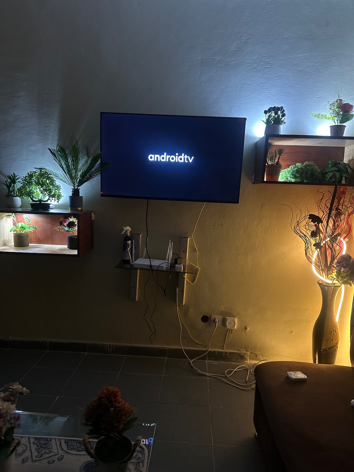 A living area is presented with a wall-mounted television displaying a logo. Surrounding shelves are filled with decorative plants, illuminated by soft lighting, enhancing the overall ambiance. A comfortable sofa is positioned nearby, with a small coffee table adorned with flowers.