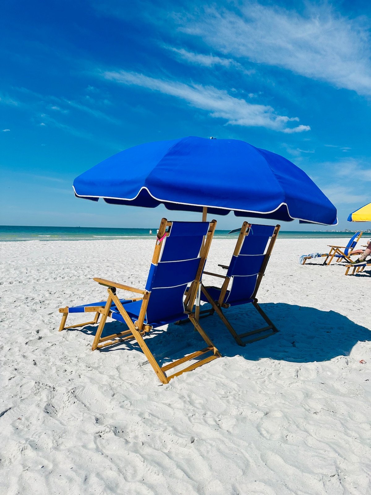 Two blue beach chairs are positioned under a large matching umbrella, resting on soft white sand. The calm ocean can be seen in the background under a clear blue sky, providing a relaxing setting for beachgoers.