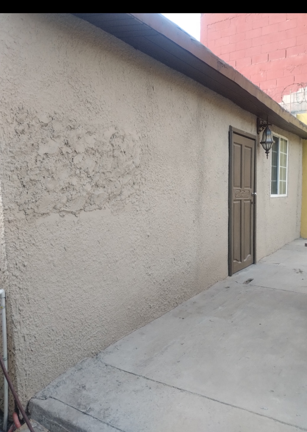 The exterior wall of a building is shown, featuring a textured surface in a neutral tone. A wooden door is located on the right side, next to a well-lit window. The adjacent concrete pathway is visible, providing access to the entrance.