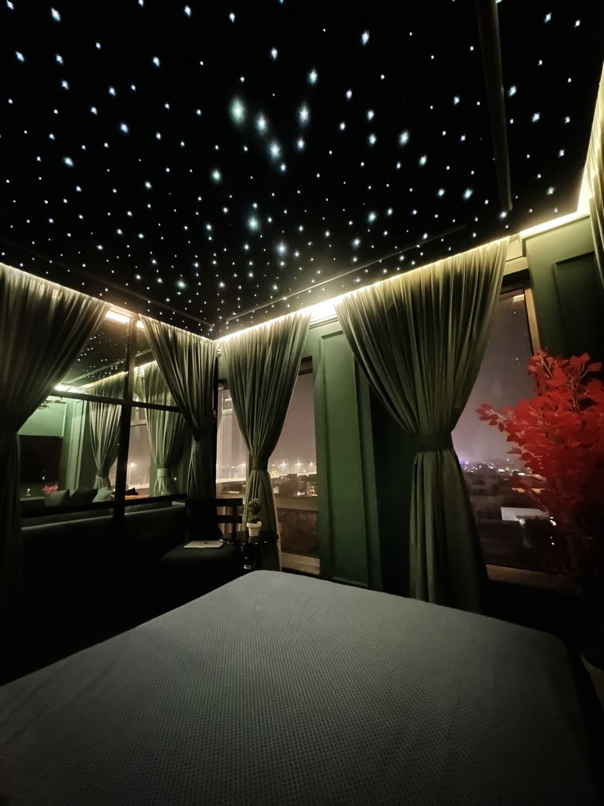 A luxurious bedroom features a star light ceiling that creates a tranquil ambiance. Large windows framed by elegant curtains offer distant city views, while soft bedding invites relaxation. A decorative plant adds a touch of warmth, contributing to the serene environment.