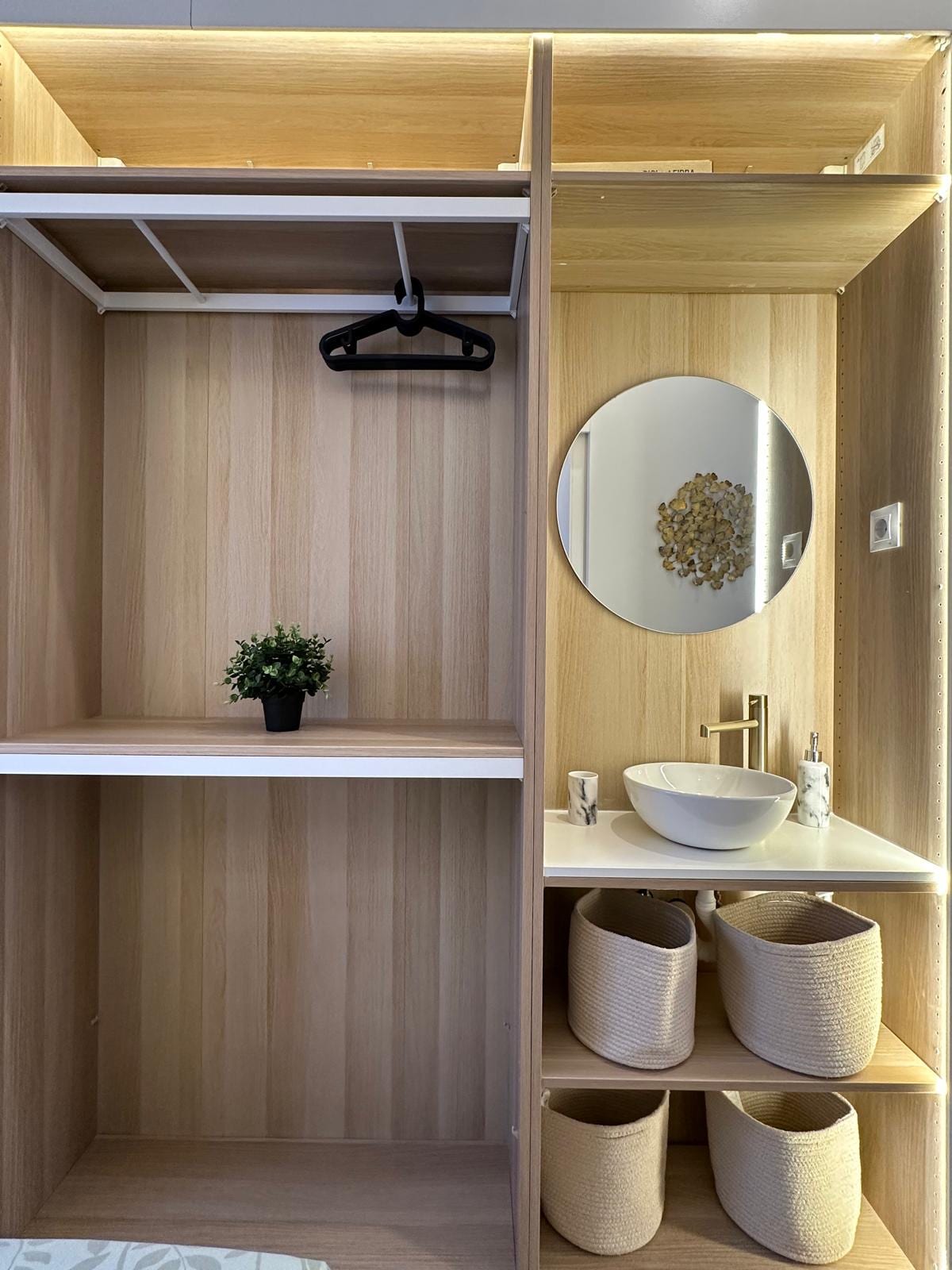 An organized wardrobe space features a hanging rod with a clothes hanger and an adjacent mirror. Below, an elegant basin is paired with a modern faucet. Three decorative baskets provide additional storage, complemented by a small plant for a touch of green.