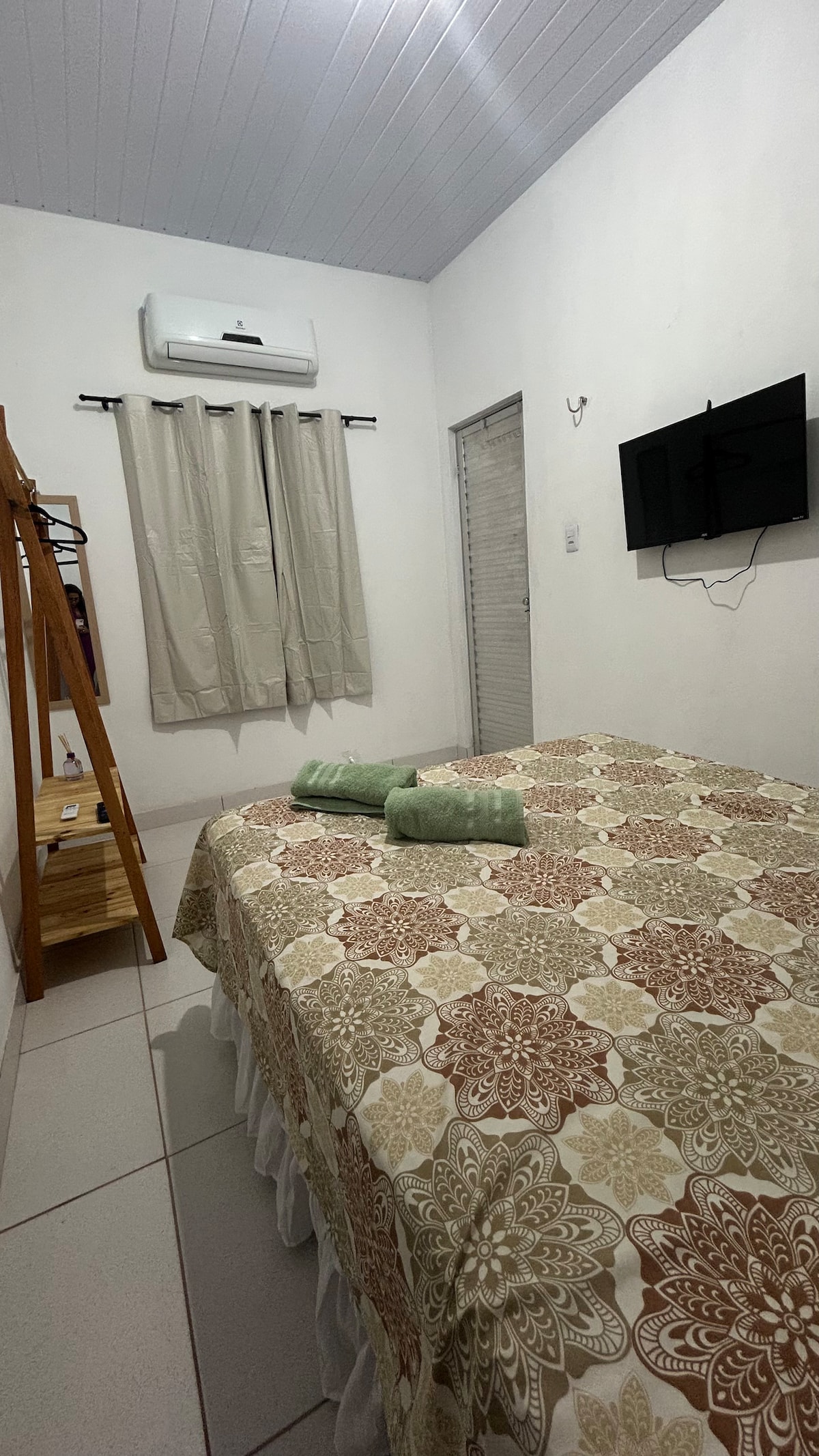 A well-organized bedroom features a comfortable bed with a patterned bedspread. Soft green towels are neatly placed on the bed. A wall-mounted television and air conditioning unit are visible. Natural light filters through light-colored curtains, creating a calm atmosphere.