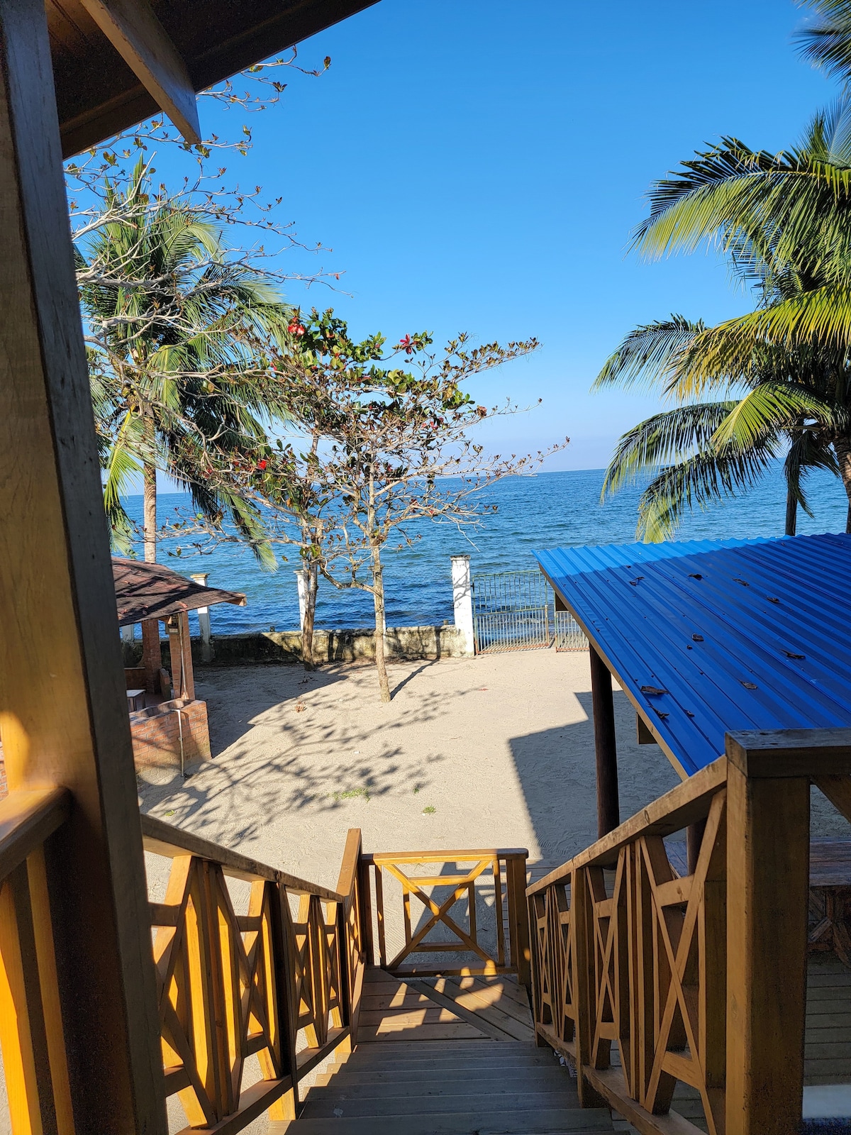 A staircase made of wood descends towards a sandy area, bordered by tropical trees. The ocean glistens in the background under a clear blue sky, while a nearby structure with a blue roof is partially visible.