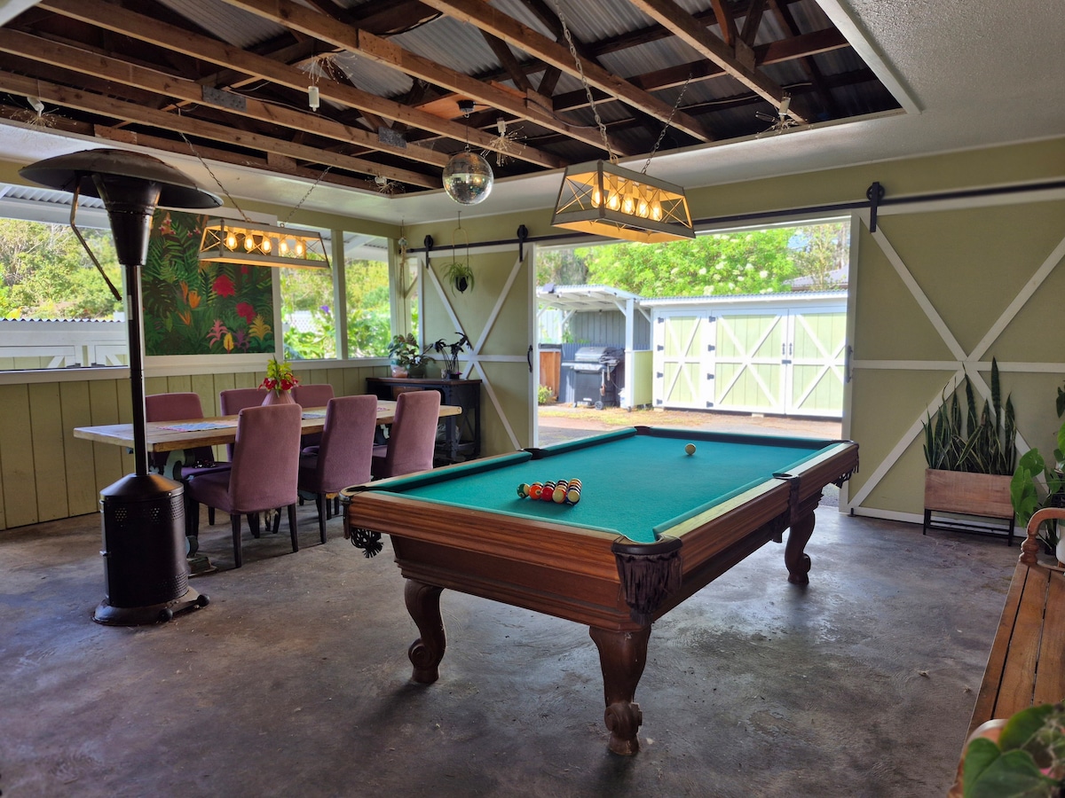 An outdoor game room features a billiard table positioned at the center, surrounded by ample natural light from large windows. A dining area with a wooden table and upholstered chairs is visible, while a heater provides warmth. Greenery can be seen through the windows.