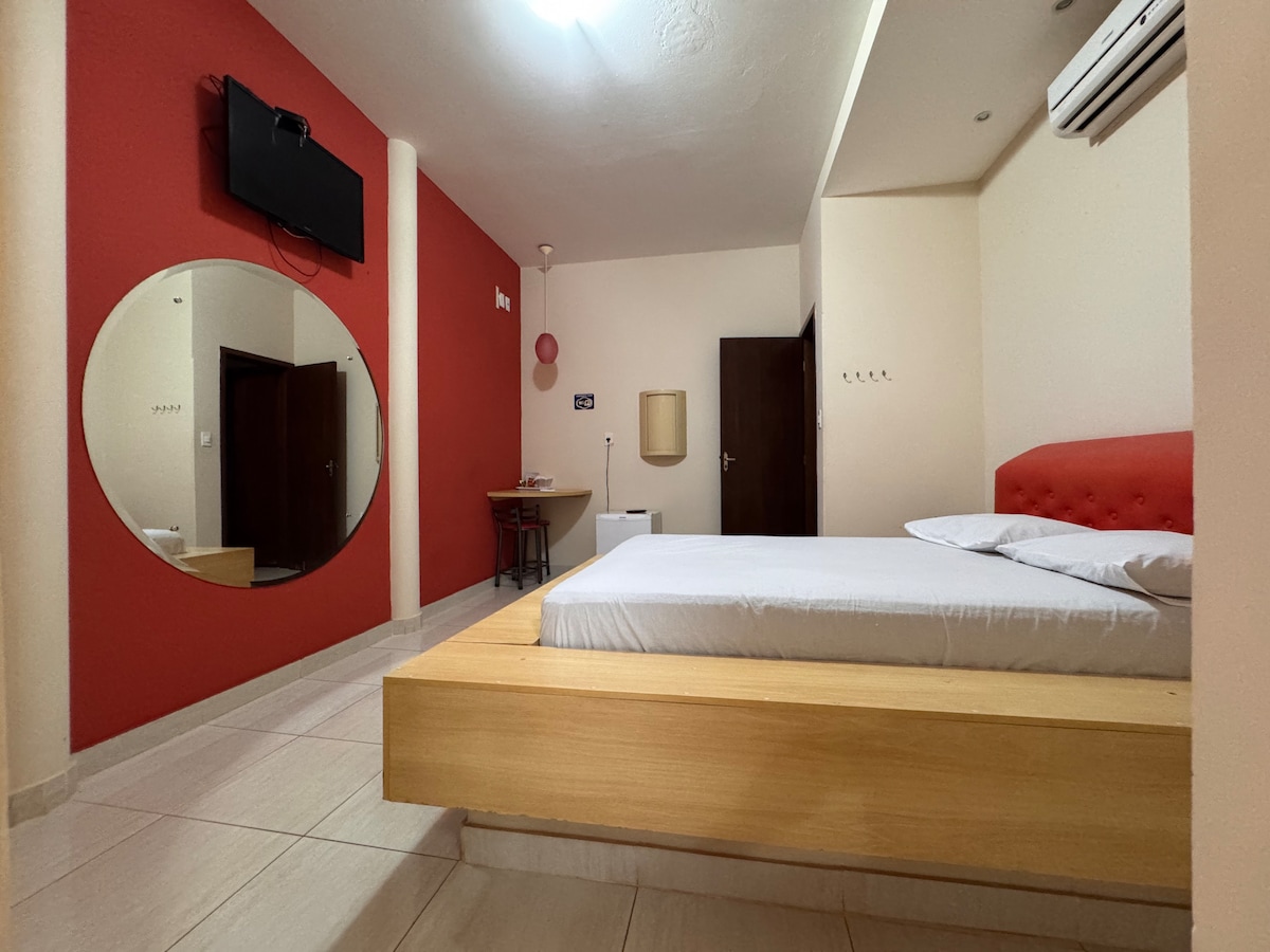 The room features a wooden platform bed with a minimalist design, complemented by a red accent wall. A round mirror and flat-screen television are mounted on the walls, while a small table is positioned beside the bed. Natural light enters through a doorway.
