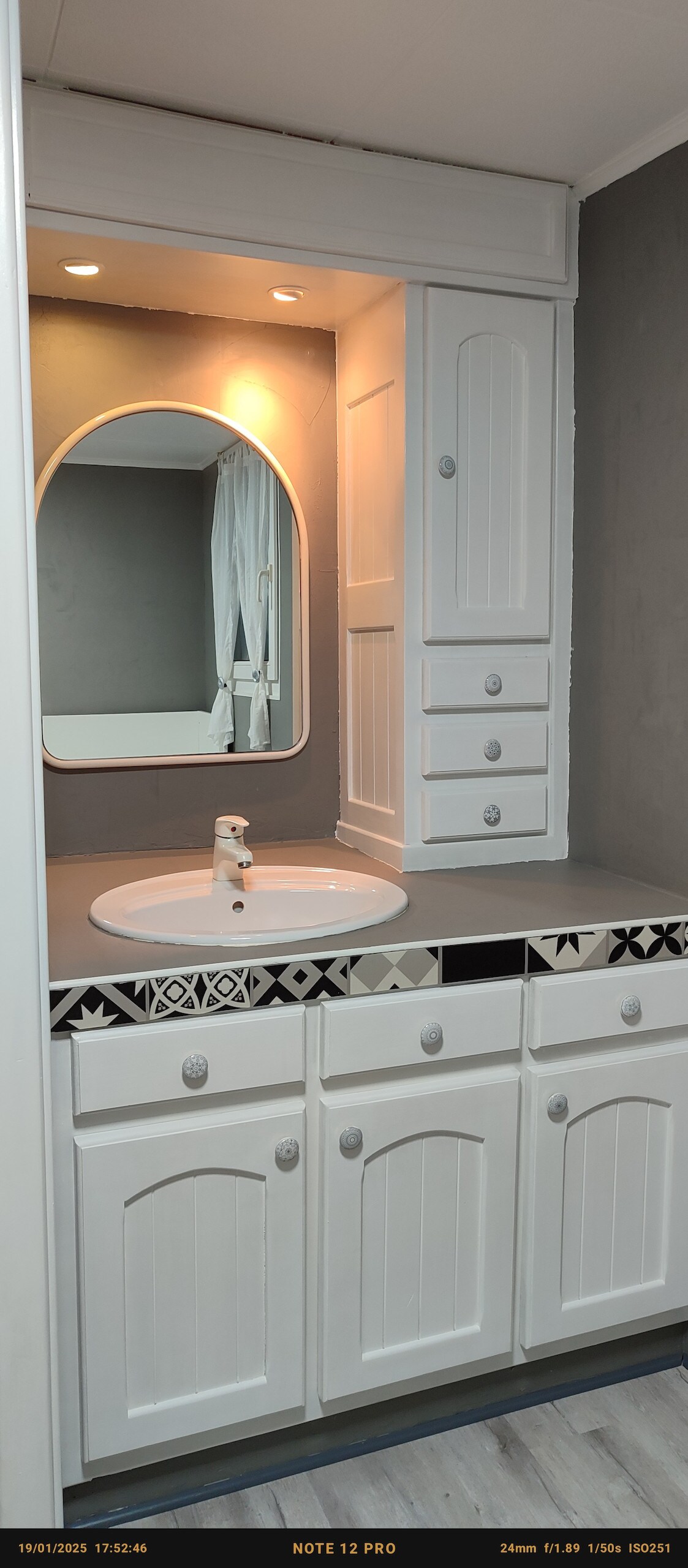 A functional bathroom area is showcased, featuring a white vanity with a round mirror above. The countertop is dark and smooth, with cabinetry providing storage space. Light fixtures illuminate the area, while a gray wall offers a subtle contrast to the white cabinetry.