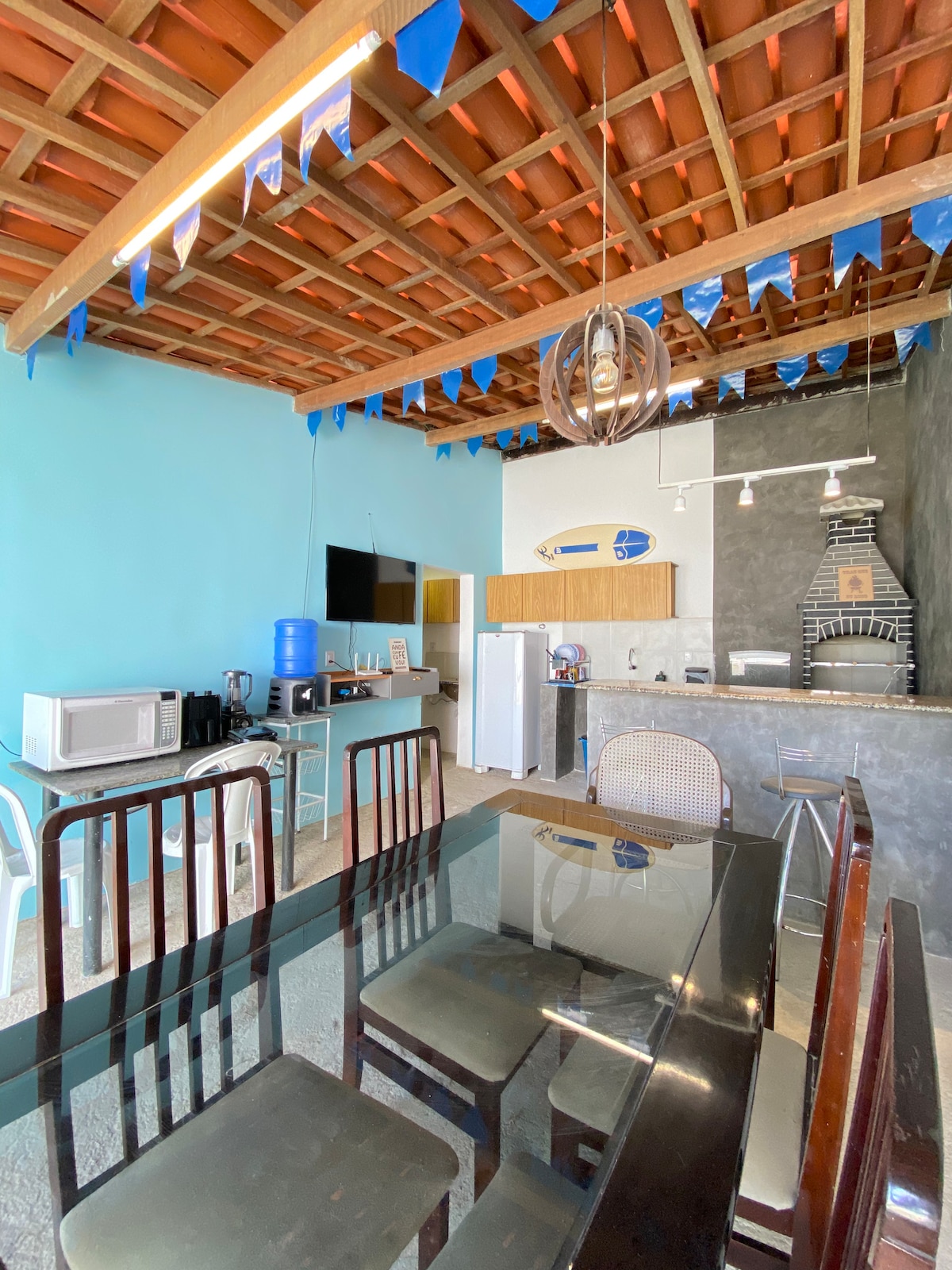 A light-filled kitchen and dining area features a glass dining table surrounded by wooden chairs. A blue accent wall complements the tiled floor. A kitchen counter with various appliances, including a microwave and a coffee maker, is visible, along with a decorative fireplace in the background.