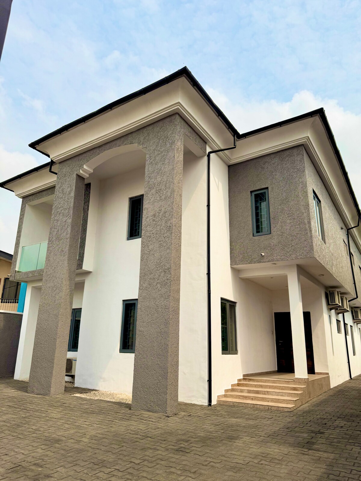 The exterior of the house is highlighted by a modern architectural design featuring a combination of white and gray walls. Large windows provide natural light, while a set of stairs leads to the entrance, framed by tall columns that enhance the structure’s elegance.