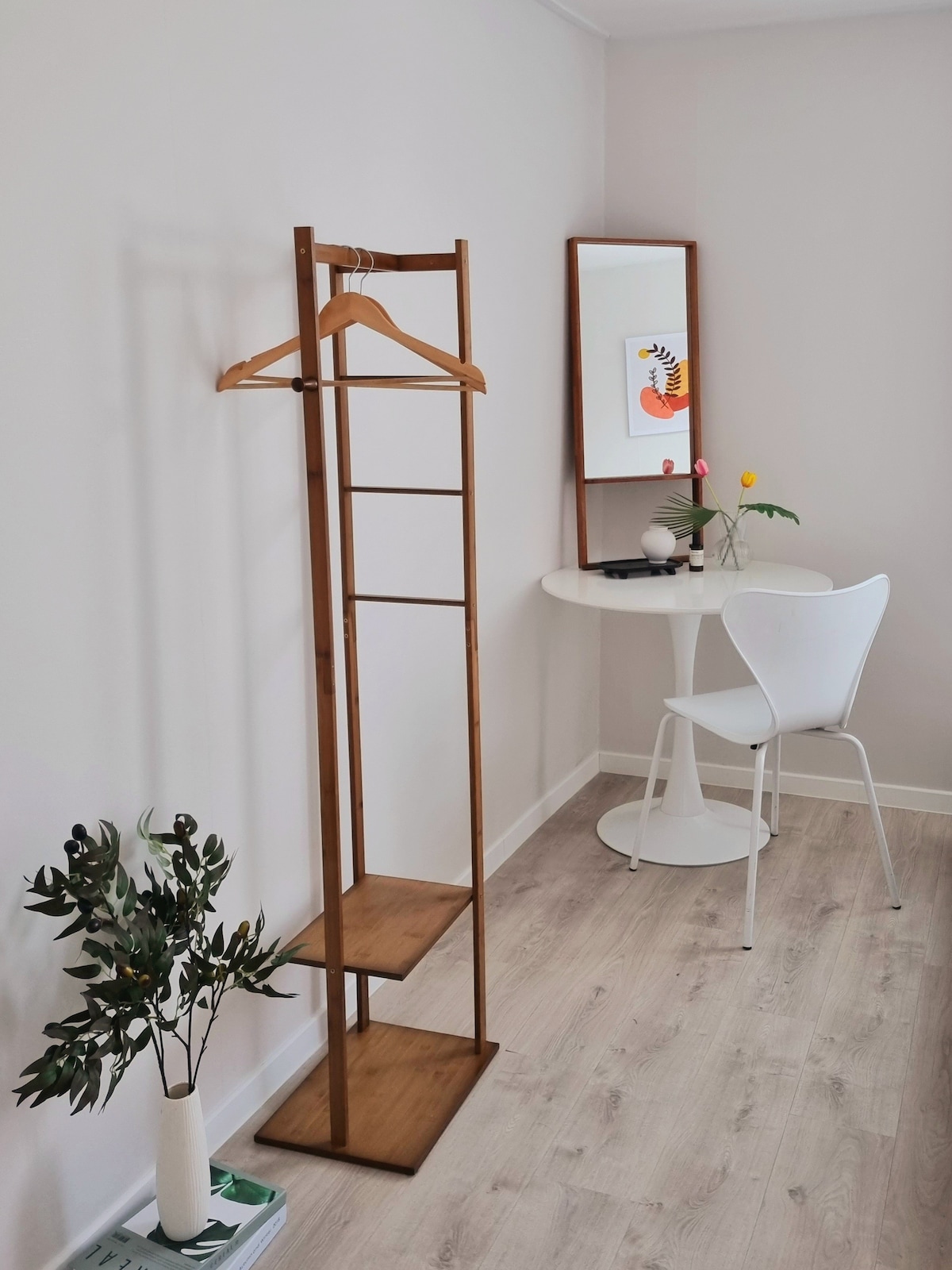 A minimalist corner features a wooden clothing rack with hangers, complemented by a small round table and a white chair. A framed artwork adds a pop of color, while a plant rests on the floor, contributing to the serene atmosphere.
