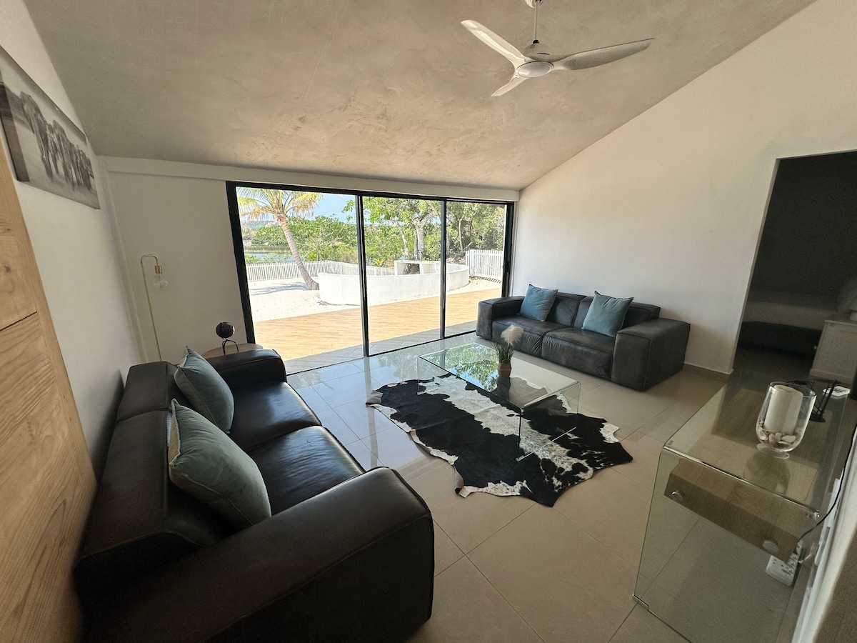 A bright living area features large glass doors, allowing natural light to fill the space. Two comfortable sofas, adorned with cushions, are positioned around a sleek glass coffee table. A minimalist design complements the serene outdoor views of the surrounding landscape.