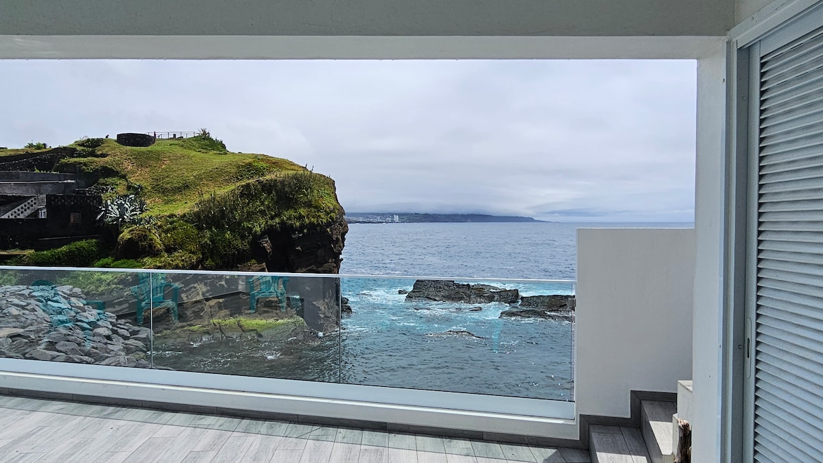 A large window frames a view of the ocean, with rocky shorelines and vibrant greenery in the foreground. The calm water meets the horizon under a softly cloudy sky, creating a serene atmosphere. A balcony railing enhances the sense of openness.