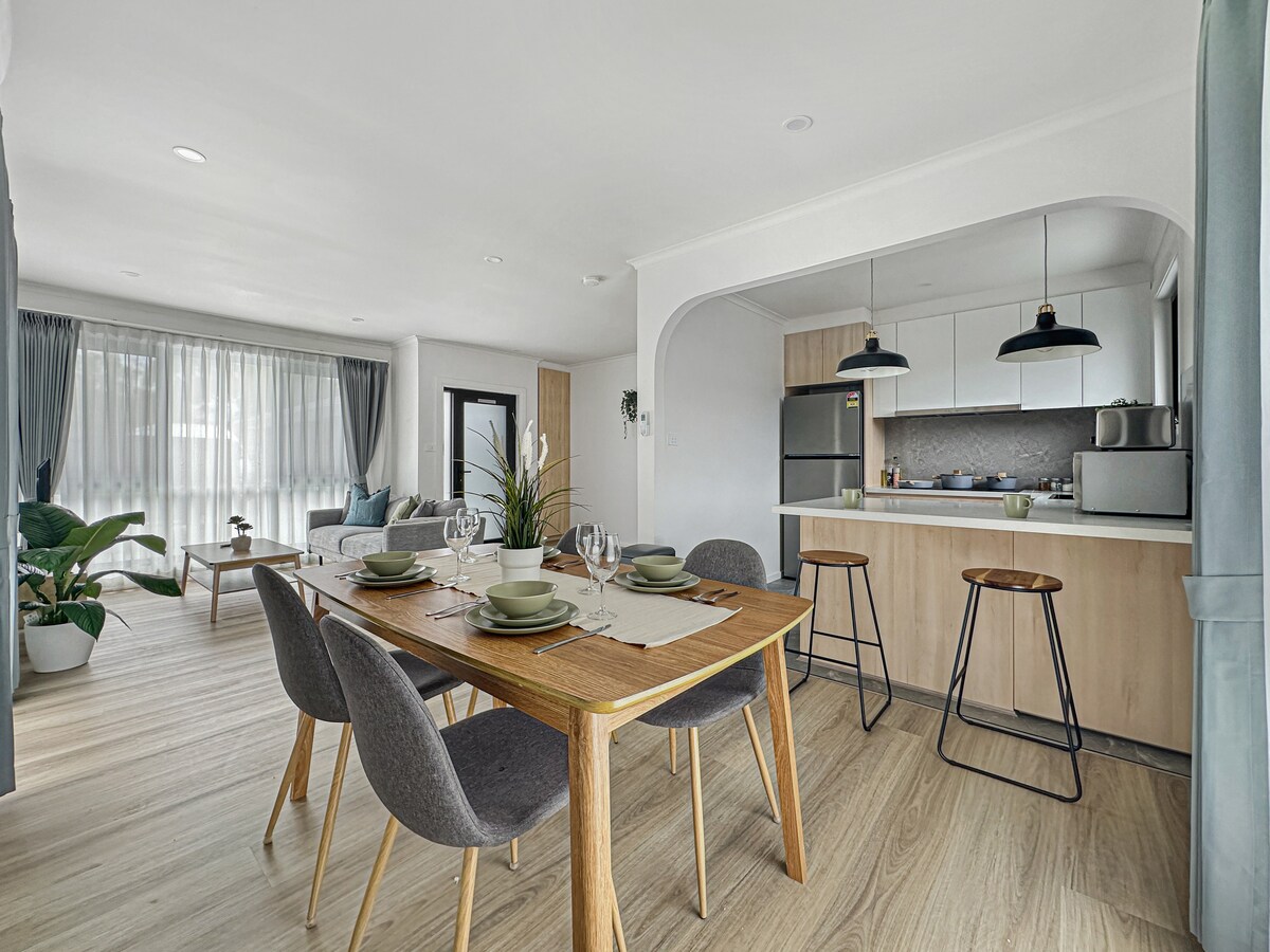 An open-plan dining area features a wooden table set for four, complemented by grey upholstered chairs. A stylish kitchen with contemporary cabinetry is visible in the background, where modern appliances and a neutral decor enhance the space's cleanliness and organization.