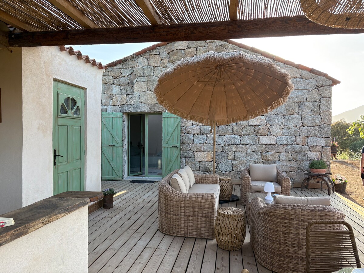 An outdoor lounge area features a thatched umbrella providing shade over comfortable wicker seating and a round coffee table. Light wooden decking leads to an exposed stone structure, with green-shuttered doors that open to the interior space.
