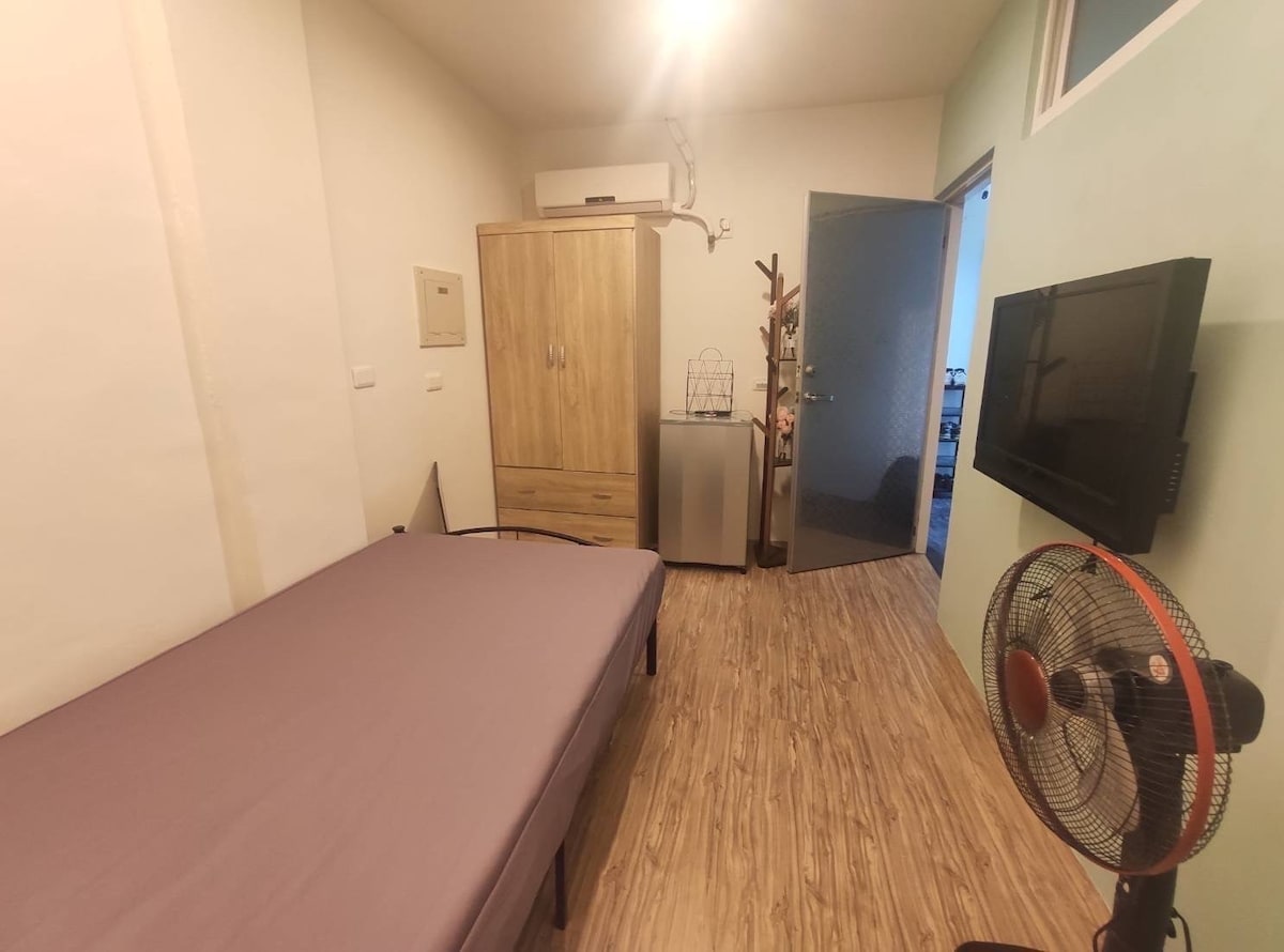 A simple bedroom features a spacious bed with a neutral-colored sheet. A wooden wardrobe and a mini-fridge are present, along with a mounted TV on the wall. Light from a ceiling fixture illuminates the room, and a fan is positioned near the bed.