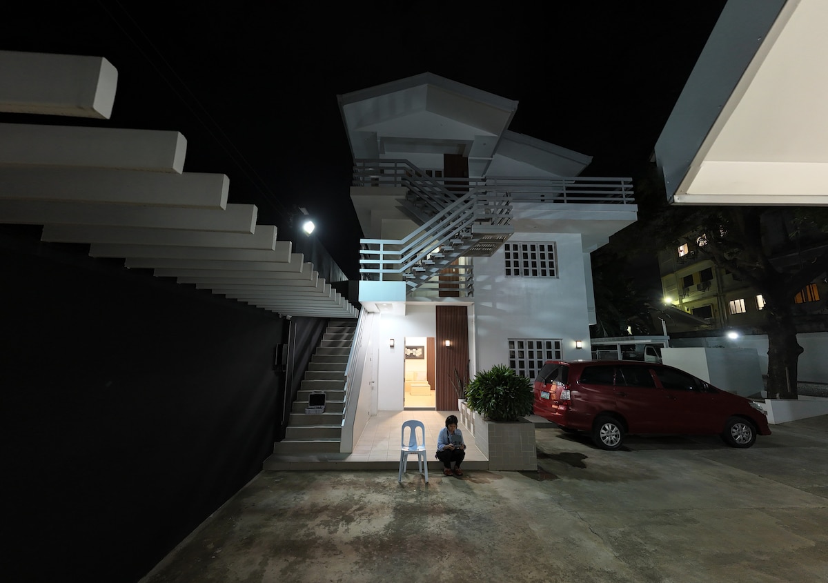 A two-story building is illuminated at night, showcasing a modern architectural style. A staircase at the front leads to the upper level. A person sits on a chair in the foreground, with a parked car and a small planter visible nearby.
