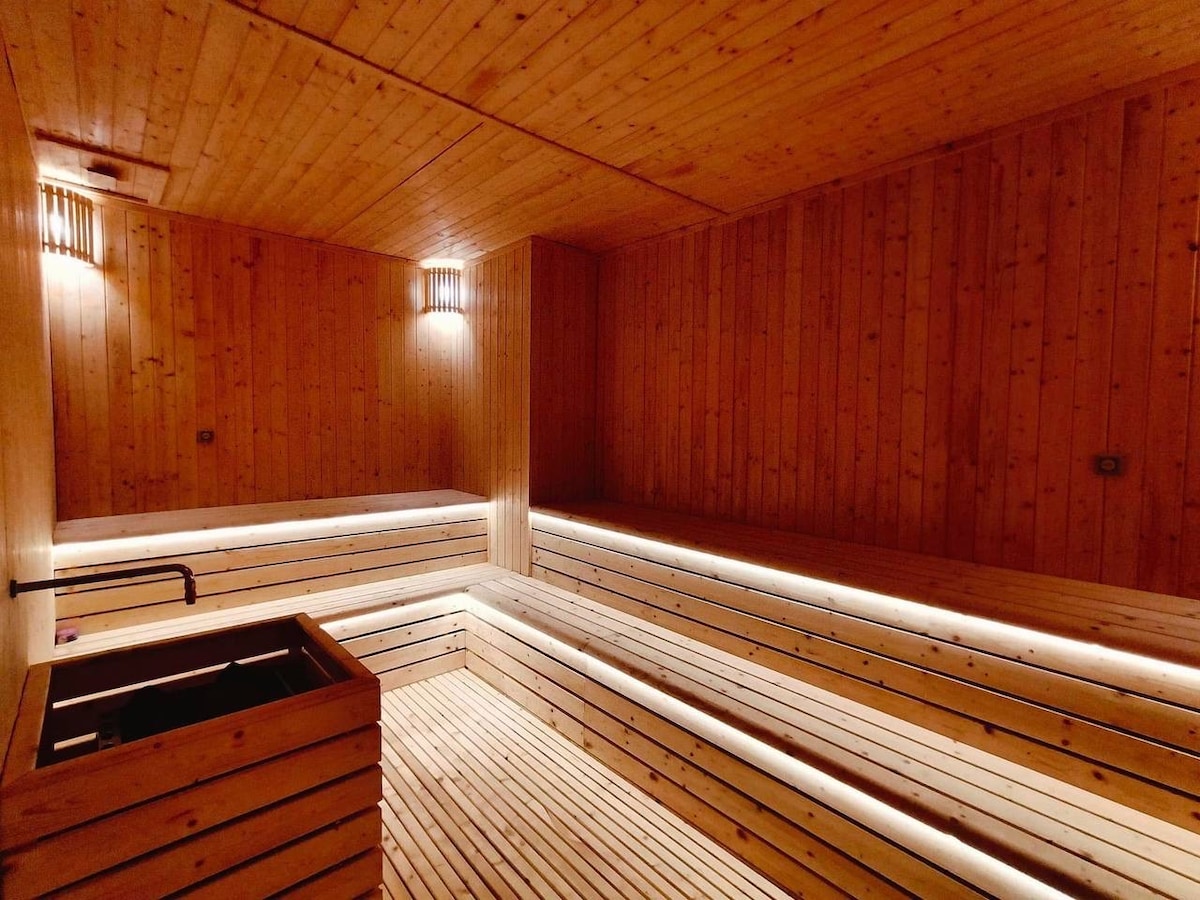A serene sauna space features warm wooden paneling and ambient lighting. Bench seating surrounds the room, providing ample seating options. A stone-lined heating element is visible, emphasizing the spa-like atmosphere in this calm and rejuvenating retreat.