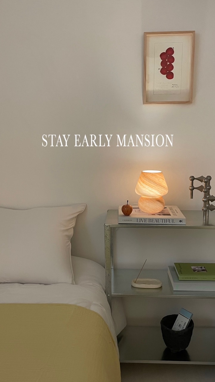 A cozy bedroom setup is depicted, featuring a soft bed with neutral bedding and a bedside table. A stylish lamp provides gentle light beside a framed artwork and various books, enhancing the calm atmosphere.