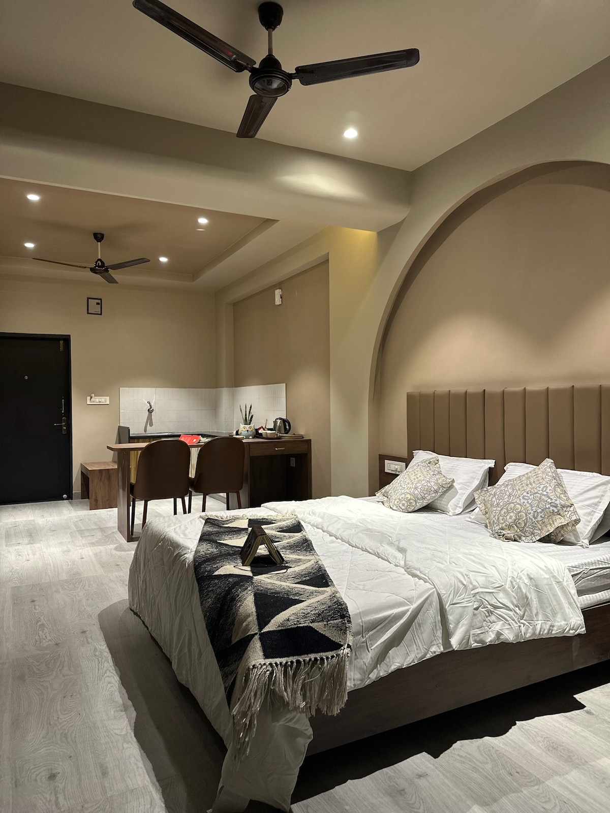 A spacious sleeping area features a large bed adorned with decorative pillows, complemented by neutral-toned walls and a cozy blanket. A dining table with two chairs is set against the wall, while the kitchenette is equipped with essential appliances, creating a functional and inviting atmosphere.