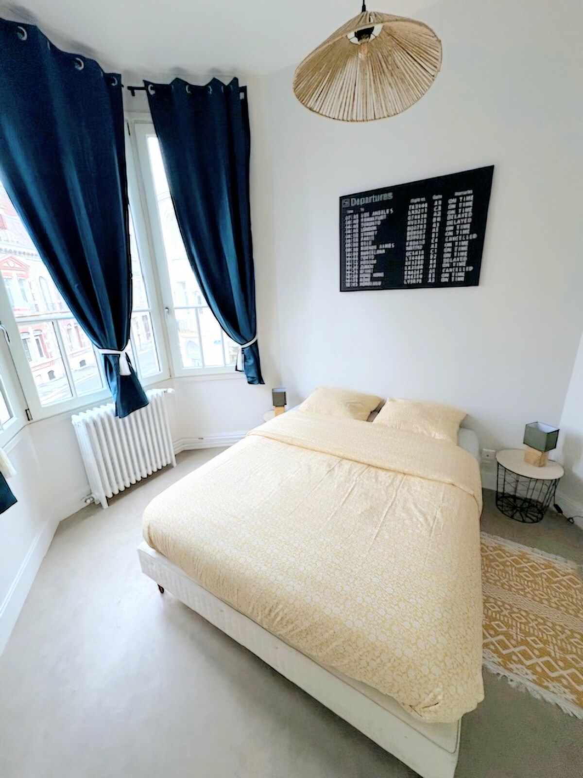 A well-lit bedroom features a spacious bed dressed in soft yellow linens. Tall windows with elegant navy blue curtains allow natural light to fill the room. A decorative wall displays a list of destinations, while a woven rug adds texture to the neutral-toned floor.