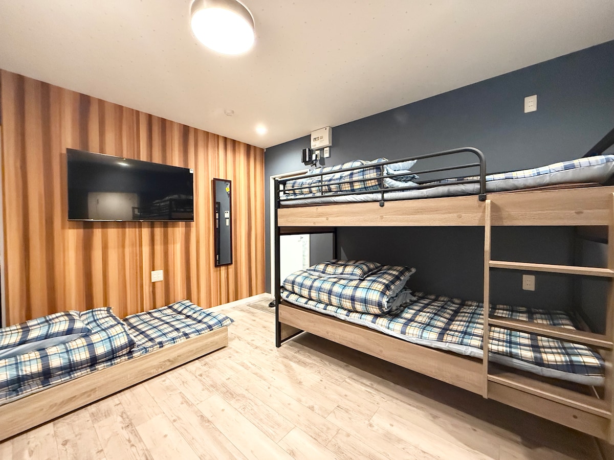 A modern sleeping area is furnished with a metal bunk bed and additional floor mattresses, all covered with plaid bedding. A flat-screen television is mounted on the wall opposite, while the room features a blend of wood paneling and soft blue walls.