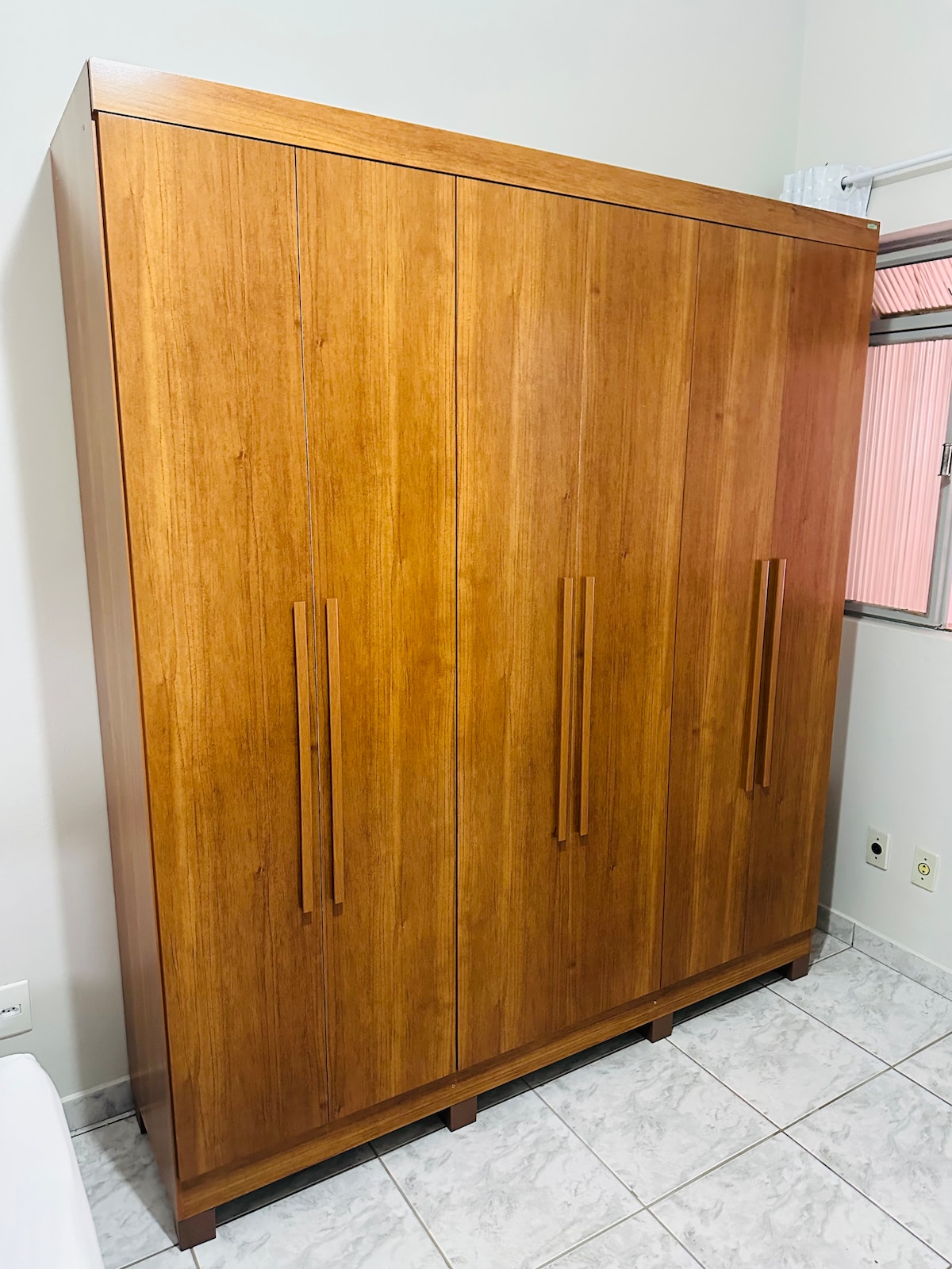 A wooden wardrobe stands against a wall, featuring a natural finish that highlights the grain. The wardrobe includes two panels with sleek handles, providing ample storage. A window is visible nearby, allowing natural light to illuminate the space.