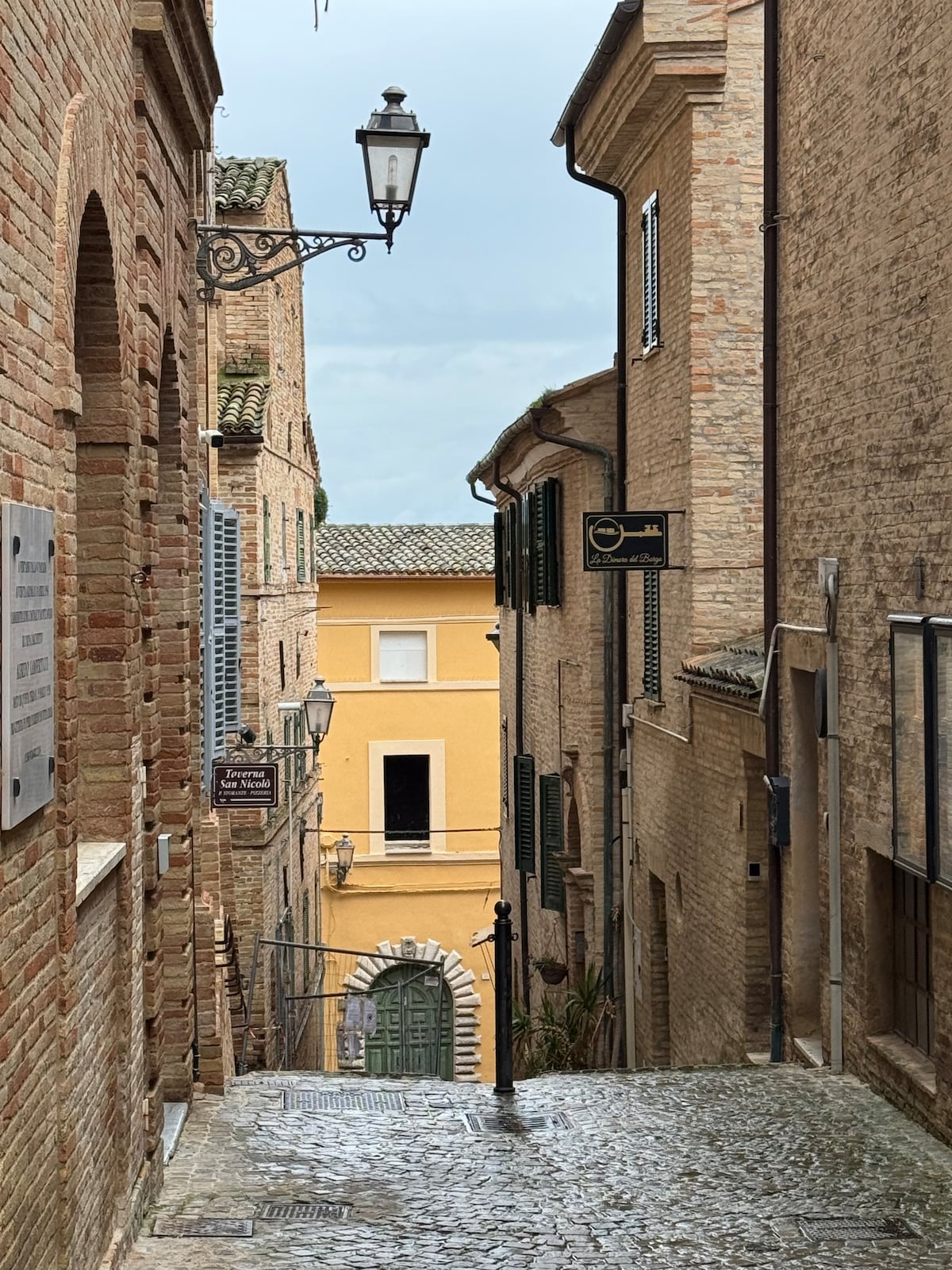 A narrow cobblestone street lined with historic buildings of warm brick tones is captured. A yellow structure with arched windows can be seen at the end of the alley, while a decorative street lamp and signage add character to the scene, evoking a charming village atmosphere.