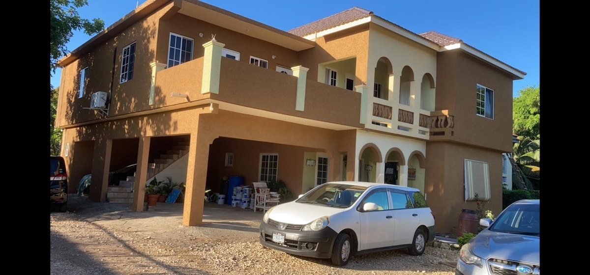 A two-story building with a warm tan exterior and balconies on the upper level. Vehicles are parked in front on a gravel driveway. Arched doorways are visible, and greenery surrounds the property, contributing to a serene atmosphere.