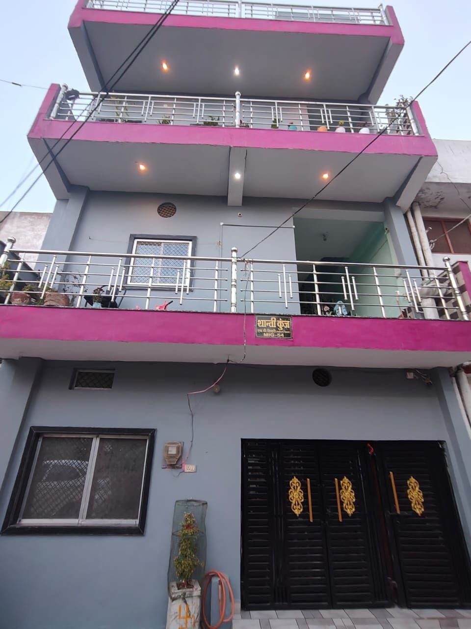 A three-story building is presented with a vibrant pink and grey facade. The entrance features decorative black doors adorned with golden handles. Balconies on the upper floors are lined with potted plants, adding greenery to the exterior. Outdoor lighting creates a welcoming ambiance in the evening.