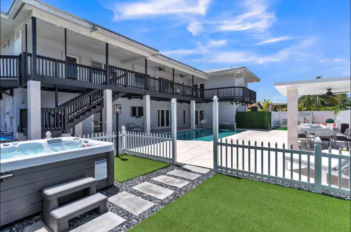 An expansive outdoor area features a sparkling pool and a hot tub, surrounded by lounge chairs and a dining space under a shaded pergola. Lush green grass enhances the space, while a decorative fence provides privacy. The two-story house with balconies adds a touch of elegance.