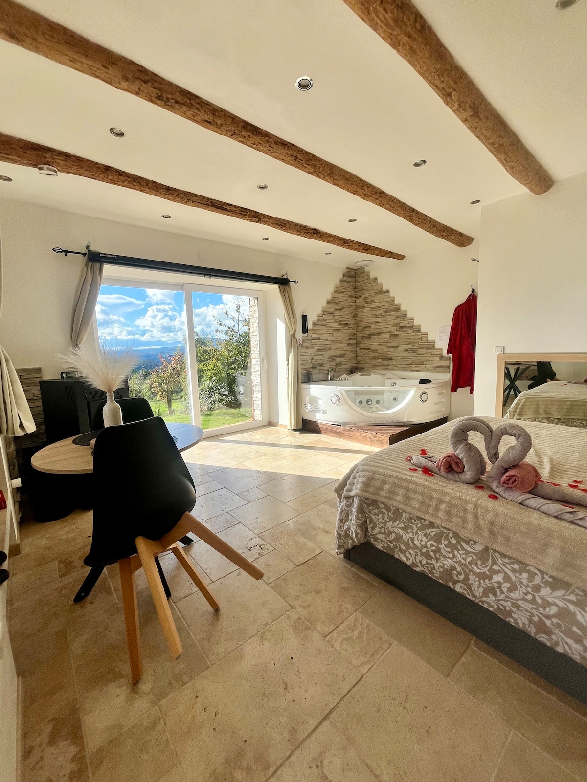 A bright studio features exposed wooden beams and stone accents. A spacious bed, adorned with decorative towels, sits beside a well-lit area. A jacuzzi is positioned near large sliding doors that offer panoramic views of the mountains and direct access to the outdoor space.