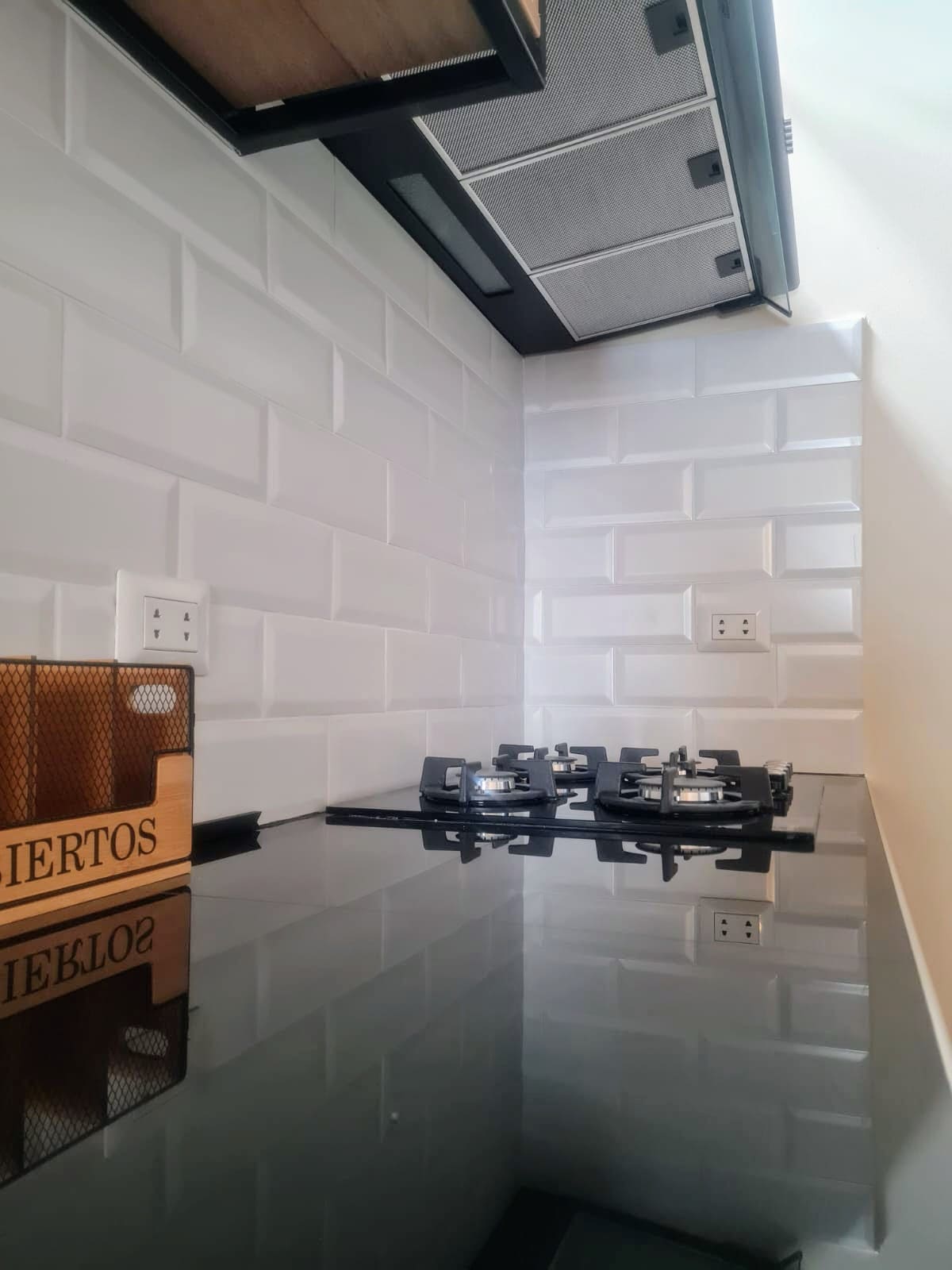 A modern kitchen area is displayed with a sleek black countertop. A gas stove with multiple burners is positioned against a textured white tile backsplash. Nearby, an organizational basket labeled 'HUECOS' is visible, adding a functional touch to the space.