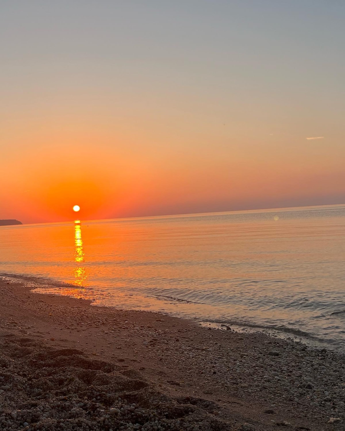 A tranquil sunset over the calm water creates a vibrant orange reflection on the surface. The shoreline, lined with pebbles, remains softly illuminated, contributing to a serene coastal atmosphere. Gentle waves lightly lap against the beach in the foreground, enhancing the peaceful scene.