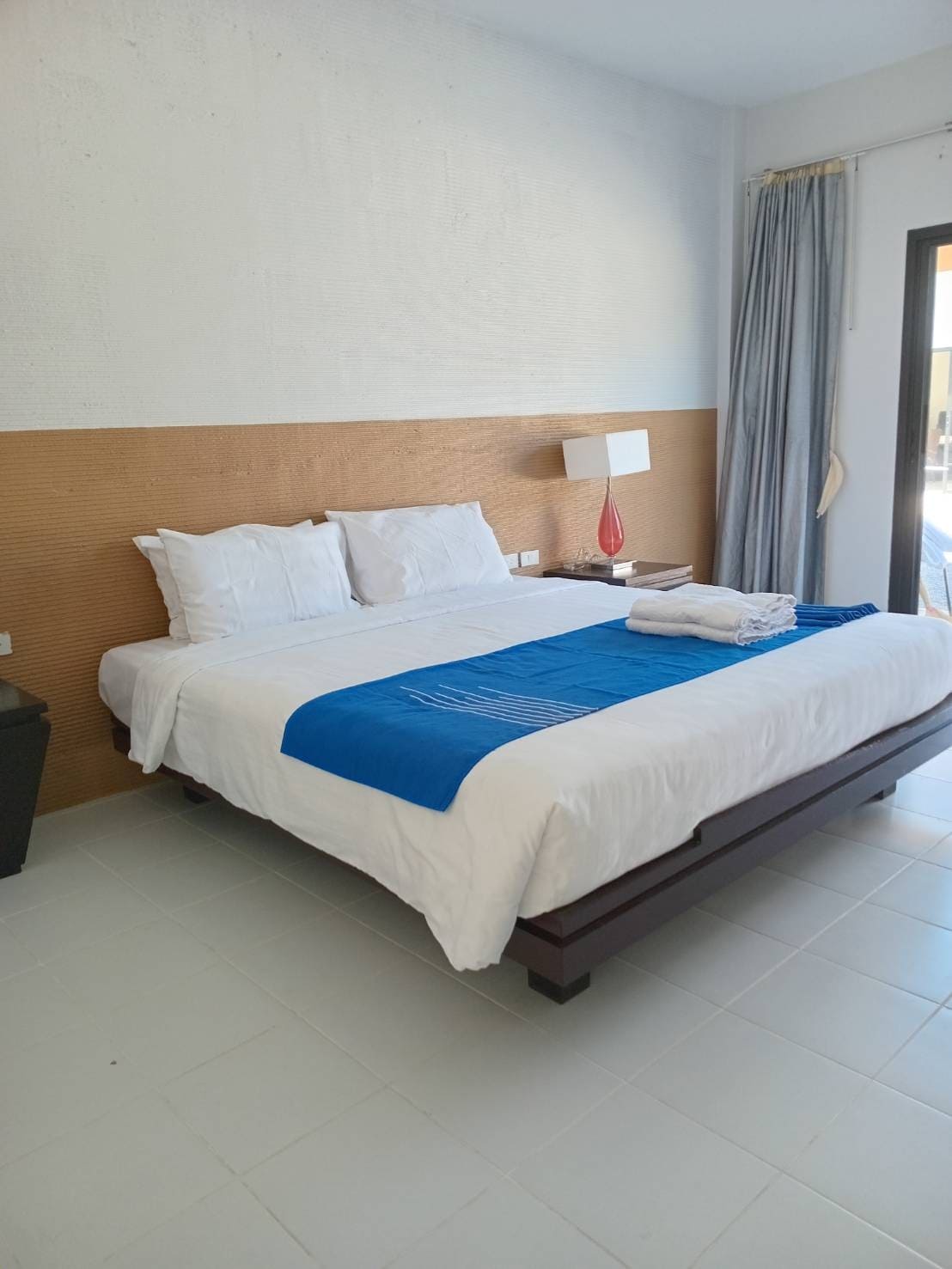A spacious bed dressed in white linens is complemented by a blue accent blanket. A small bedside lamp is positioned on a nightstand, while light drapes gently at a window, adding a bright touch to the room's neutral palette.