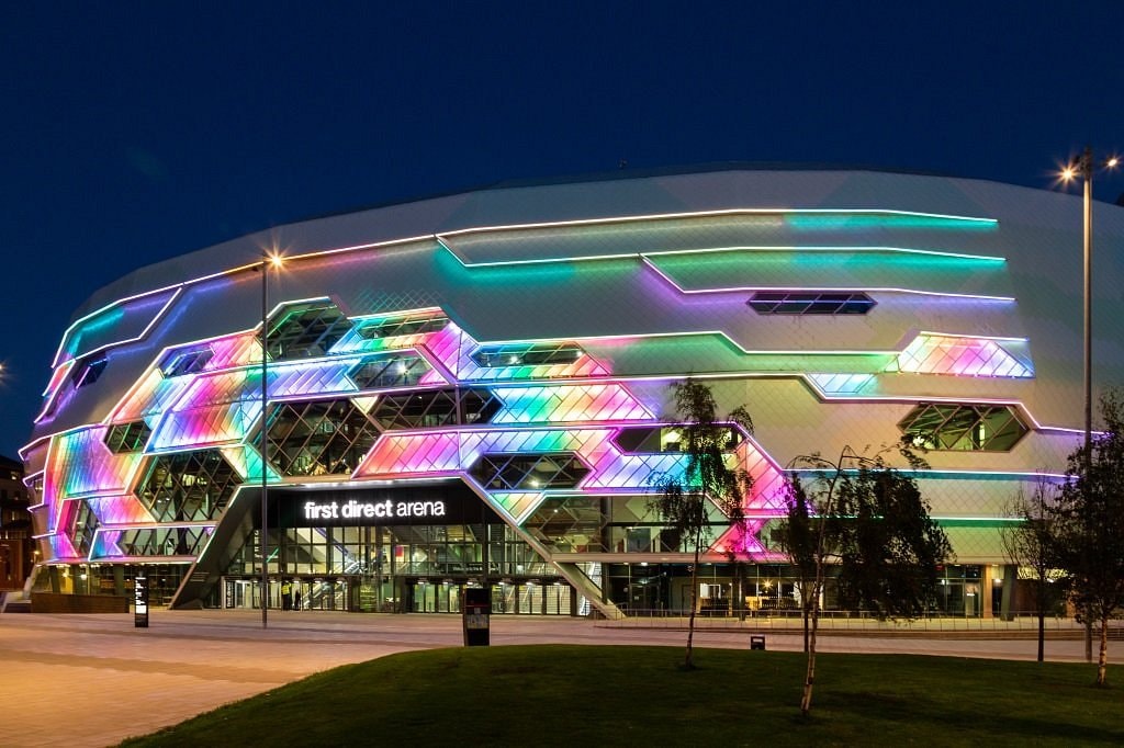 The First Direct Arena is illuminated at night with vibrant, colorful lights across its modern architectural facade. The building features a geometric design with large glass panels reflecting the surroundings, creating a dynamic visual effect. A landscaped area with trees is visible in the foreground.