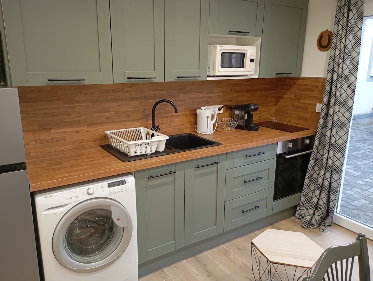 A modern kitchen is presented with cabinetry in a muted green tone. Essential appliances include a washer, microwave, coffee machine, and an under-counter refrigerator. A wooden countertop complements the space, with a sink and dish rack positioned thoughtfully. A small table and chair offer a dining option.
