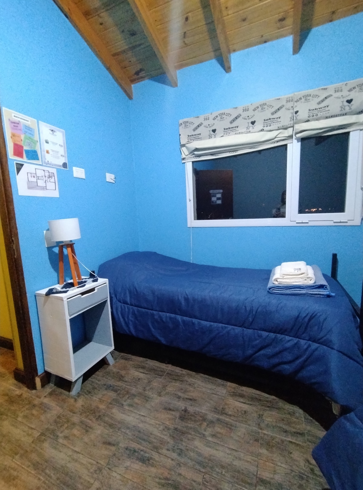 A bright blue-walled room features a single bed covered with a dark blue blanket. A bedside table with a modern lamp sits beside the bed. Natural light enters through a well-dressed window, and informative materials are visible on the wall.