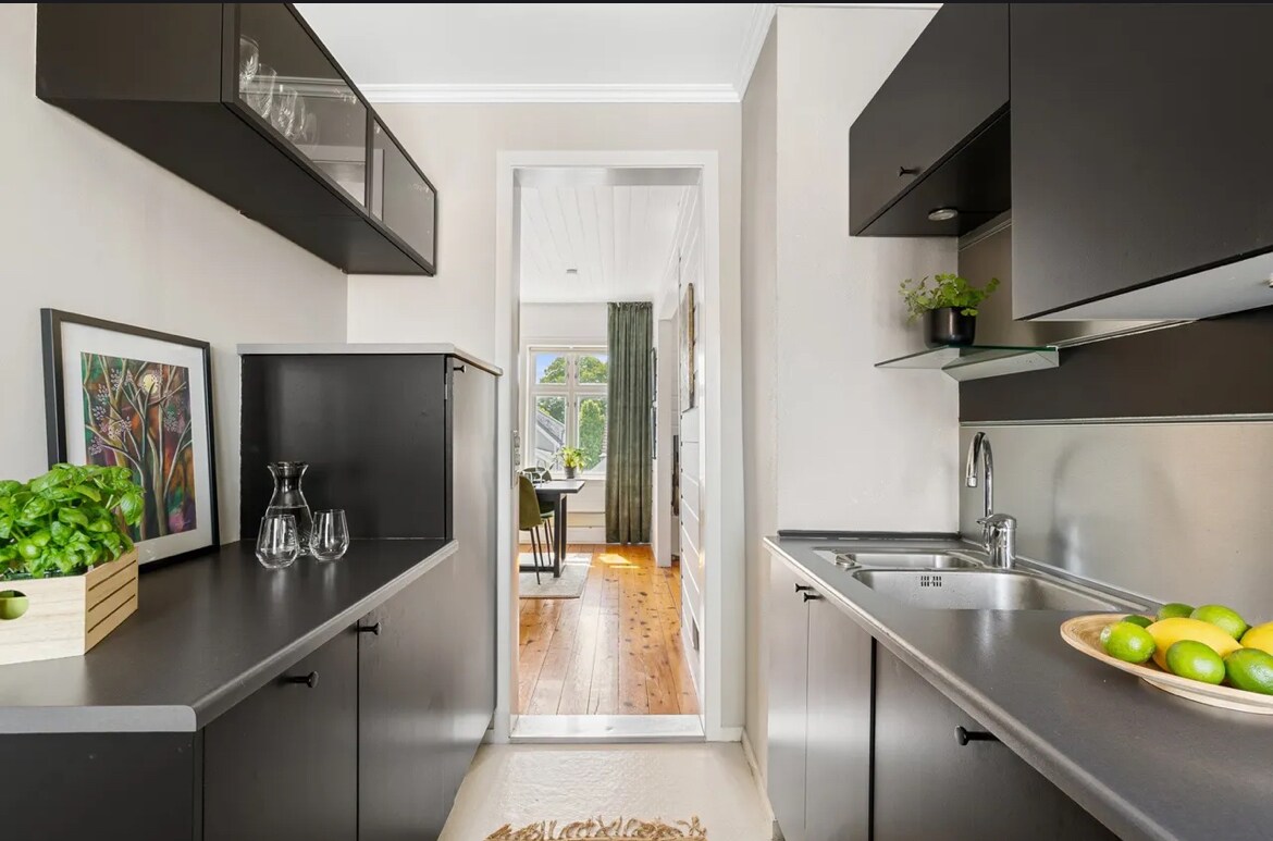 The kitchen area features dark cabinetry and a sleek countertop. An abundance of natural light is provided through a nearby window. A bowl of green apples is placed on the counter, and plants are visible on the shelves, promoting a fresh atmosphere.