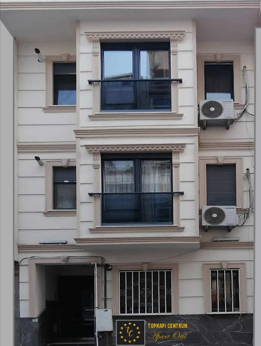 The exterior of the building displays a modern architectural style, featuring three evenly spaced windows on the upper levels. Air conditioning units are mounted next to the windows, and a secured entrance is visible at the ground level, providing access to the property.