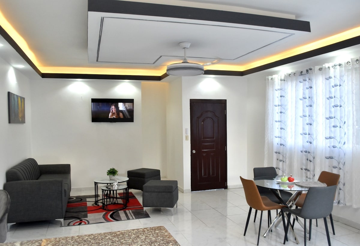 The living area is designed with modern furnishings, including a gray sofa set and a glass dining table for four. A flat-screen TV is mounted on the wall, and large windows allow natural light to fill the space, complemented by sheer curtains.
