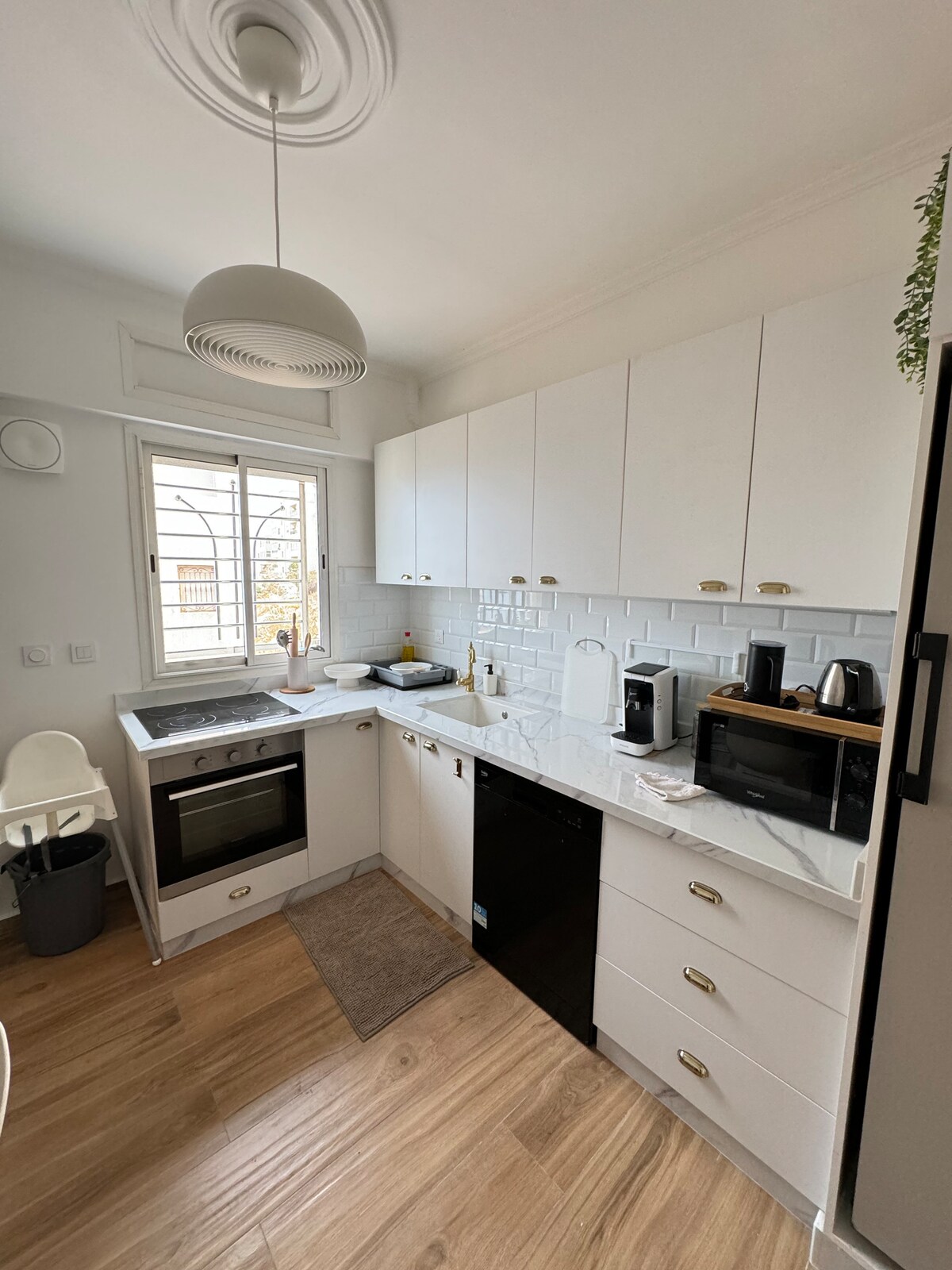 A modern kitchen features white cabinetry equipped with gold hardware. A marble countertop complements the built-in appliances, including an oven and a microwave. Natural light filters through a window, illuminating the space, while a small rug adds a touch of warmth to the tiled floor.