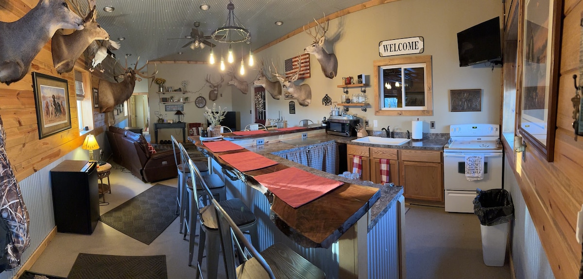 A spacious kitchen area is highlighted by a long countertop with bar seating. Decorative taxidermy deer heads adorn the walls, complemented by rustic wooden accents. The kitchen features modern appliances, a television, and a cozy seating area with a sofa, creating a comfortable gathering space.