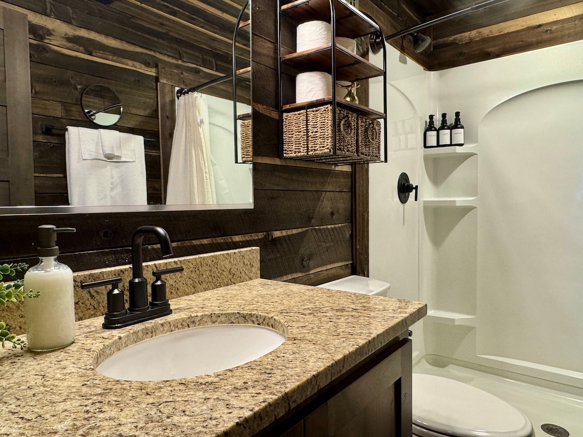 The bathroom features a granite countertop with a circular sink, complemented by dark fixtures. Shelving above holds neatly arranged toiletries and towels. A walk-in shower is enclosed with smooth white walls, enhancing the space's cleanliness. Natural wood accents add warmth to the overall design.