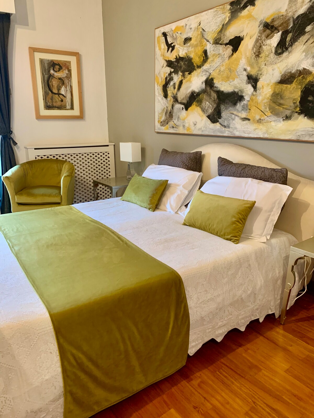 A serene sleeping area features a spacious bed adorned with decorative pillows and a smooth green throw. An armchair with a soft texture complements the room's design. Natural light filters through the window, while an abstract art piece adds visual interest to the wall.