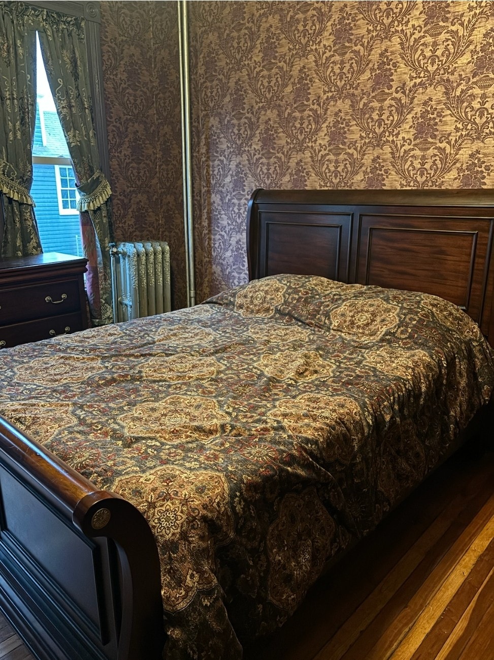 A warmly decorated bedroom is highlighted by a queen size bed covered with an ornate blanket. Richly patterned wallpaper complements the wooden furniture, including a dresser. A window with ornate curtains allows natural light to enhance the space, while a nearby radiator adds a vintage touch.