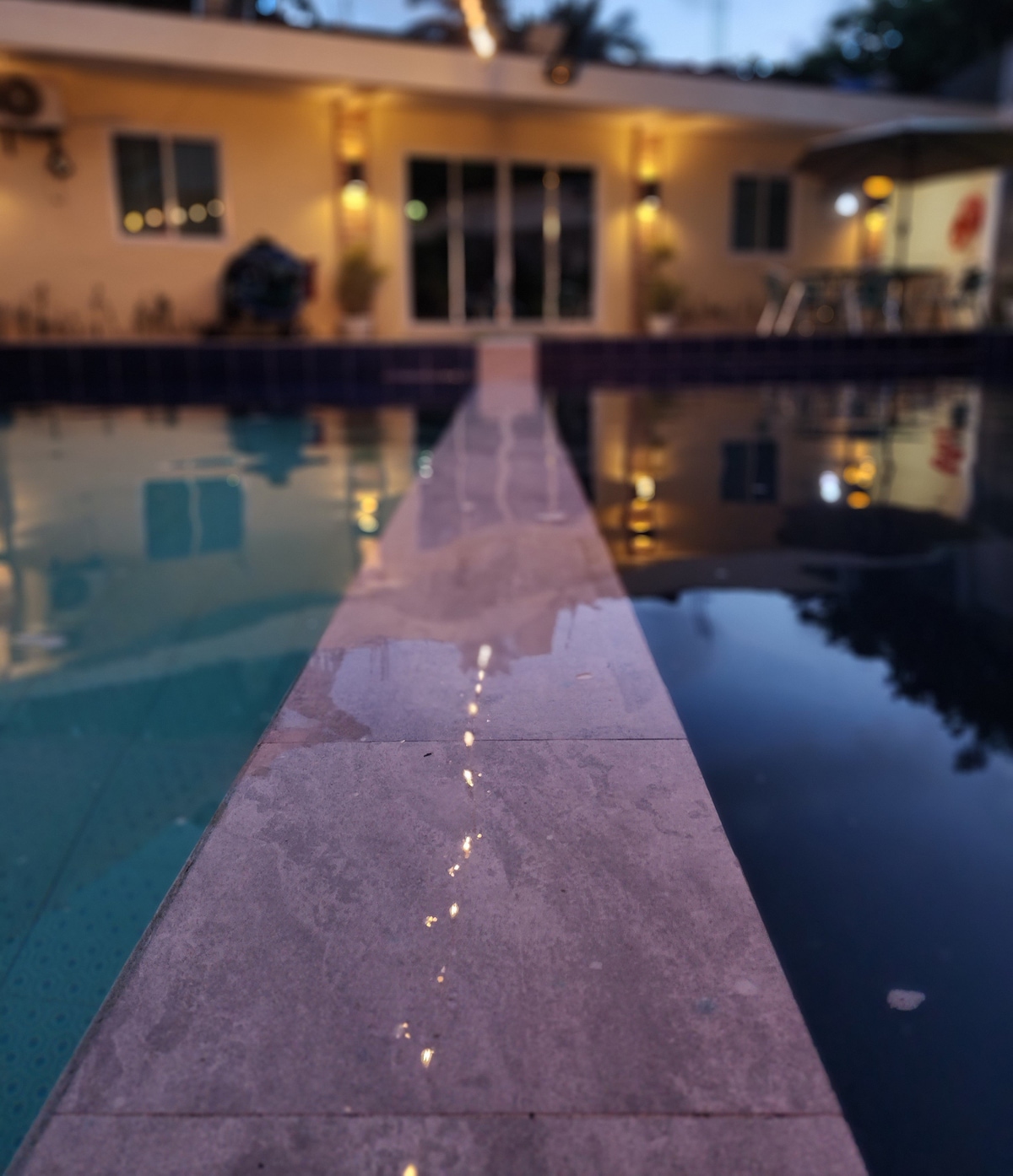 A walkway lined with smooth stone extends between two swimming pools. The area is illuminated by ambient lighting, highlighting the water's calm surface. Nearby, a patio can be seen with tables and chairs, creating a serene setting for relaxation.