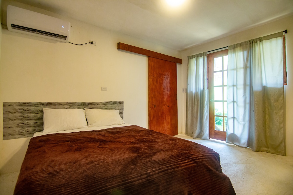 A comfortable sleeping area features a spacious bed with a soft brown blanket. Natural light filters through a set of glass doors, which open to a small outdoor space, while curtains add a gentle touch to the room.