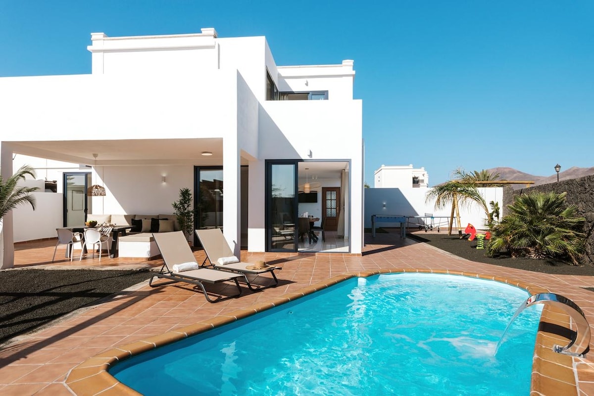 An inviting outdoor area features a private, heated pool surrounded by sandy tiles. Sun loungers provide space for relaxation. The structure consists of a two-story building with large glass doors, allowing natural light to flow into the living spaces. Lush greenery is present throughout the yard.