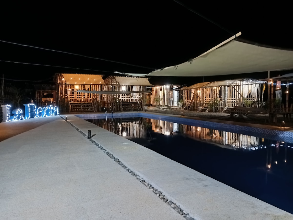 A tranquil outdoor area is illuminated at night, featuring a large swimming pool reflecting lights from surrounding structures. A wooden deck and tents create a cozy atmosphere, while the name 'La Berra' is softly lit near the pool, highlighting the relaxing ambiance.