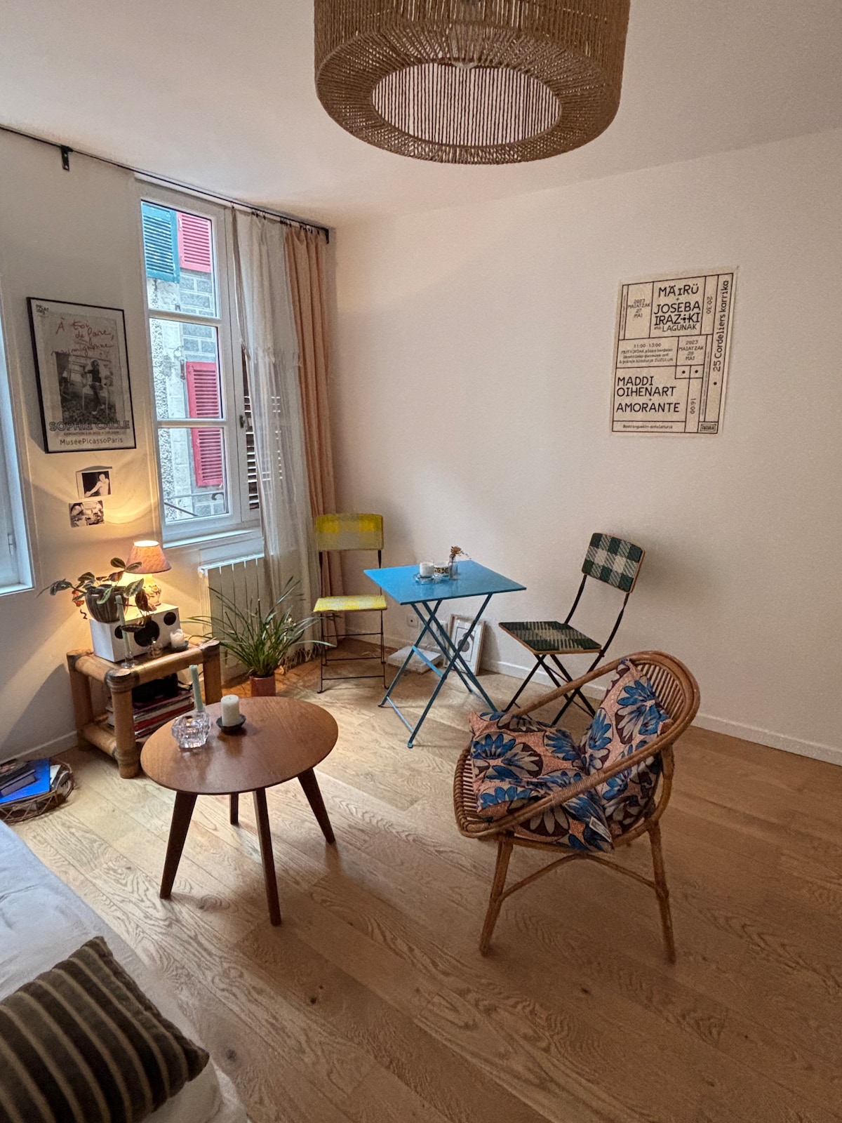 A bright living space features a light wood floor and neutral walls. A round pendant light hangs above a small blue table with two colorful chairs. A comfortable rattan chair displays a floral cushion, and decorative plants and artwork adorn the area.